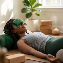 A woman lies on a yoga mat in a bright room, wearing a green sleep eye mask and a gray tank top with green leggings. She rests on a green pillow with yoga blocks and a diffuser in the background.