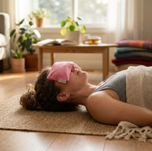 A woman lying on a woven rug in a bright living room, wearing a pink eye mask and a beige knit blanket. The mask has a small white tag with the brand name wobble