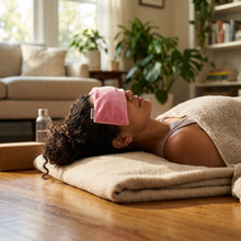 A woman lies on a wooden floor in a bright living room using a pink velvet eye pillow and wrapped in a beige blanket home relaxation