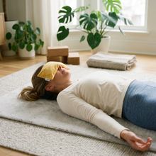 A woman lies on a light gray textured rug in a bright home room, wearing a Turmeric Velvet Washable Rectangular Eye Pillow
