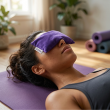 A woman lying on a purple yoga mat with her eyes covered by a purple fabric eye mask, in a softly lit indoor space with rolled exercise mats and plants in the background.