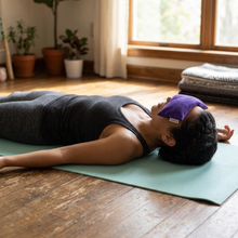 A person lies on a yoga mat in a sunlit room, wearing a purple eye mask. The person is dressed in a dark gray tank top and leggings, with potted plants, a window, and a cushioned floor visible in the background.