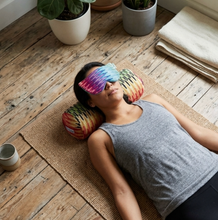 A woman lies on a yoga mat on a rustic wooden floor, wearing a colorful rainbow-patterned eye mask and resting her head on two matching rainbow neck pillows. She is wearing a gray tank top.