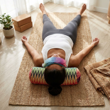 A woman lies face down on a woven yoga mat in a bright room, wearing a colorful rainbow tie on her forehead and a patterned cylindrical pillow under her head. She is dressed in a white tank top and gray leggings, with yoga blocks and a small potted plant nearby.